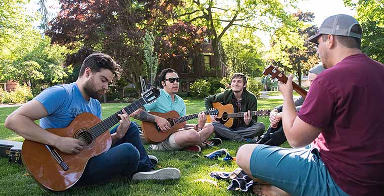 5 Students sitting on the grass at campus playing guitar