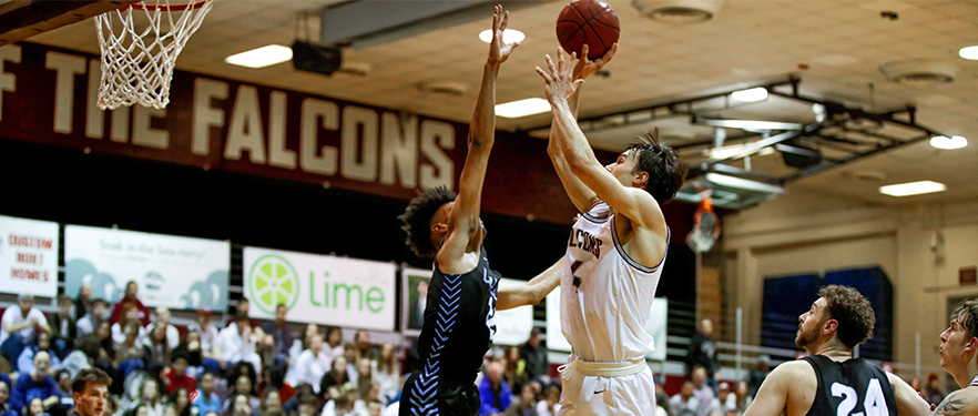 An SPU men's basketball player shoots into the hoop while an opposing player goes up for a block.