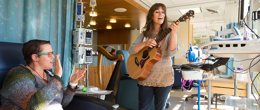 An SPU music therapy alum plays her guitar while a patient claps.