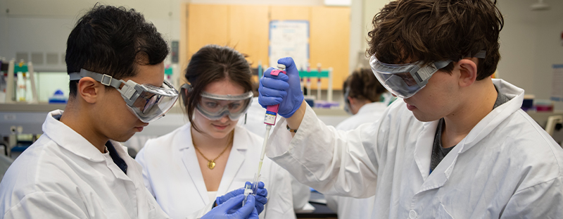 Three SPU students wearing white lab coats, protective goggles, and blue exam gloves work together in a lab. 