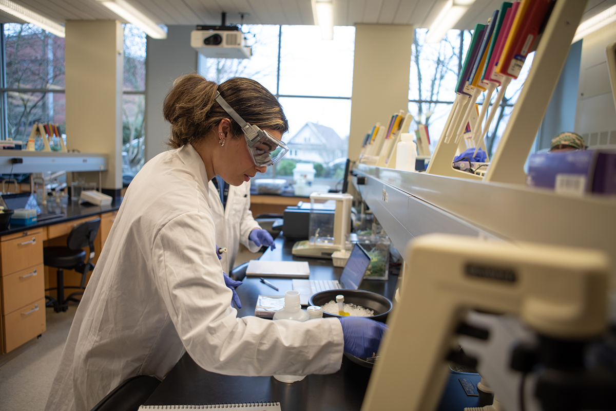 A female SPU student, wearing a white lab coat and protective googles, works in a chemistry lab