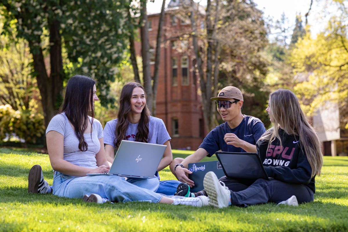 Four SPU students, three woman and one man, sit in Tiffany Loop during the spring, talking and laptops on their laps.