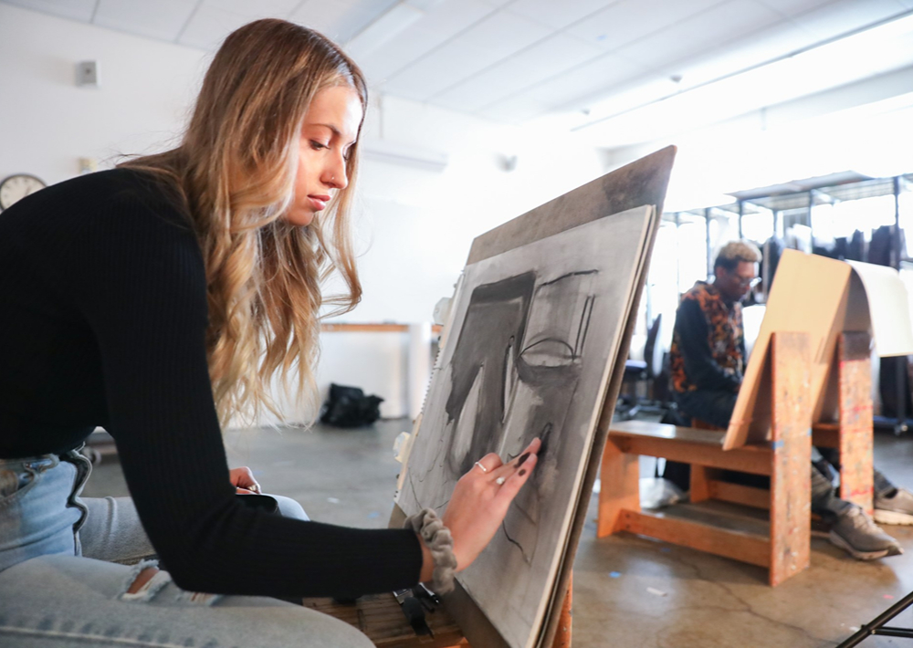 A female student in the foreground and a male student near a window both work on their charcoal drawings in drawing class at Seattle Pacific University