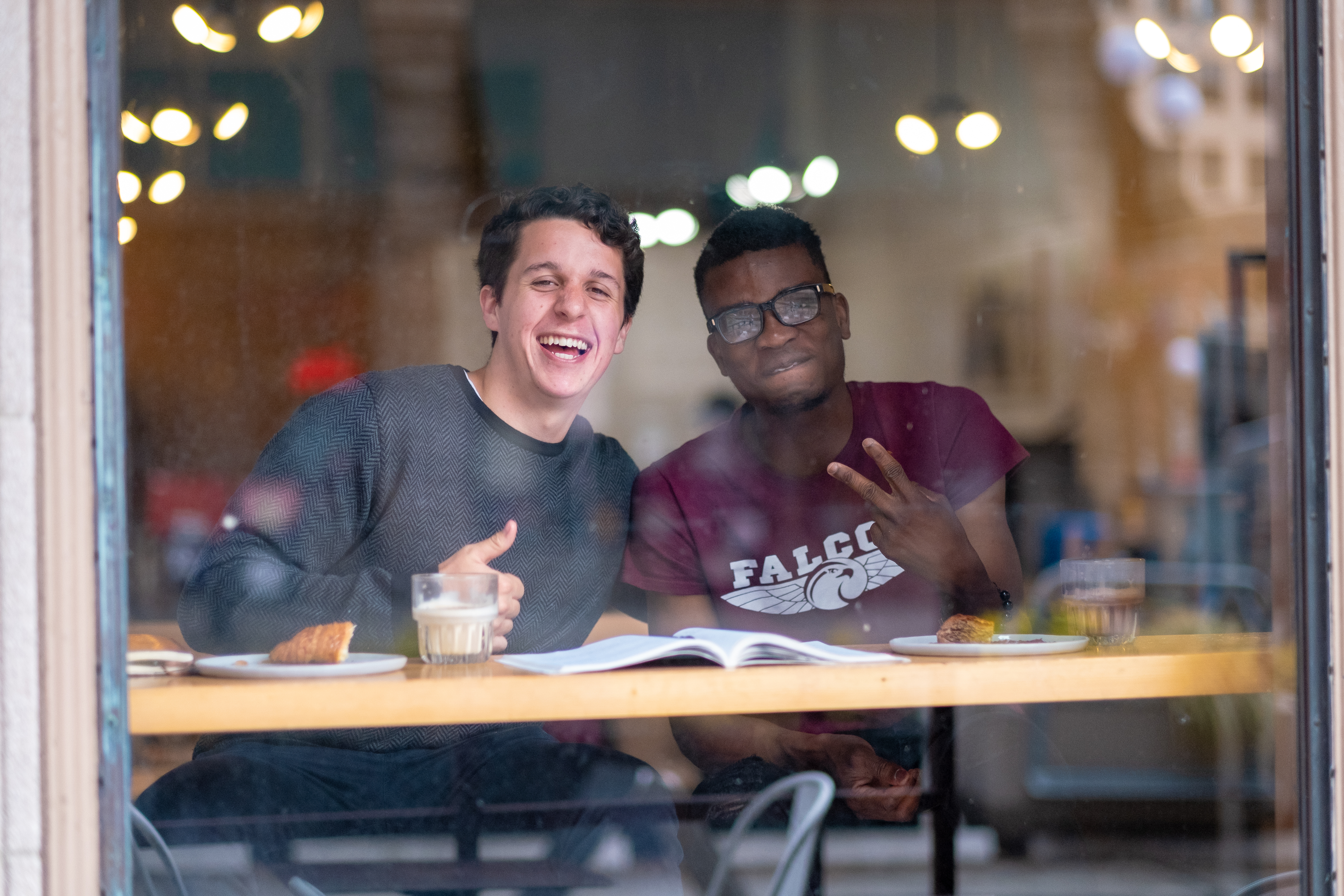 Two students smiling at the camera through a glass window.