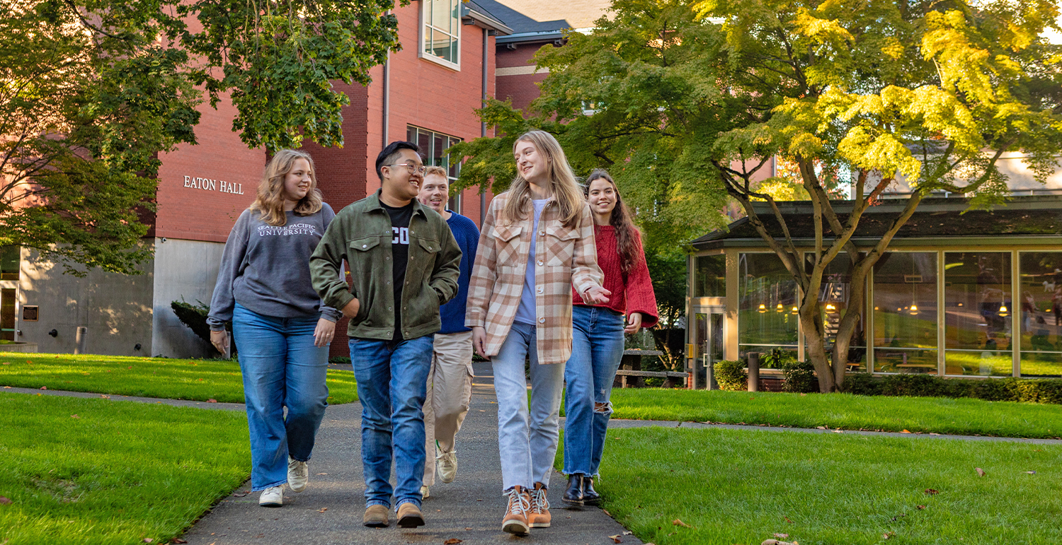 Five SPU students walking in SPU's Tiffany Loop, with the Student Union Building and Eaton Hall in the background.