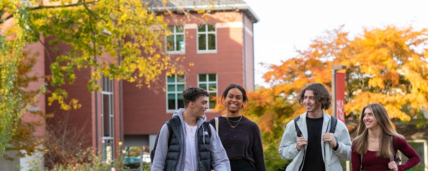 Four SPU students, three male students and a female student, walking on campus with the Eaton Science Building in the background.