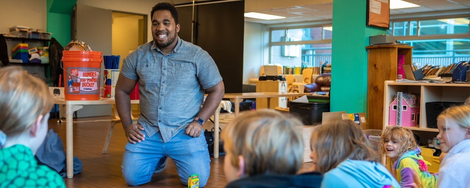 A male education student smiles as he kneels on the floor in front of young children.