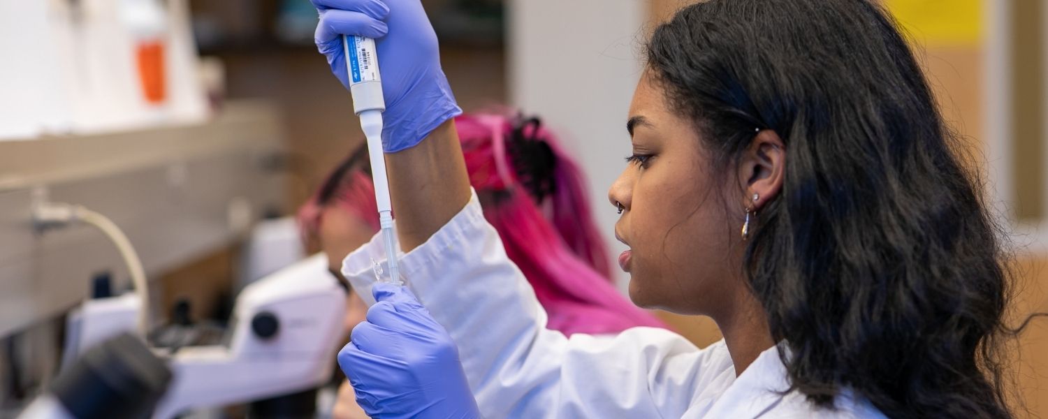 An SPU student works in a lab, adding a liquid to a test tube, as her fellow student works in the background.