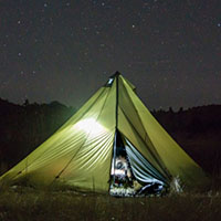 An illuminated tent sits in a dark meadow under a starry sky