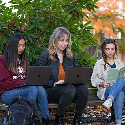 Three women students sitting on benches in Tiffany Loop while working on their laptops.