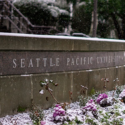 A wintertime image of crocuses dusted with snow in front of a decorative wall engraved with Seattle Pacific University's name located off Martin Square