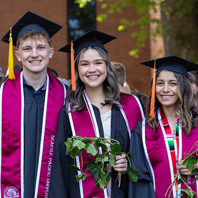 Three SPU graduates in their graduation caps and gowns, including a male student on the far left, and two female students beside him, all holding ivy from the annual Ivy Cutting Ceremony at the beginning of Commencement weekend