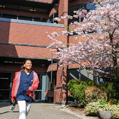 An SPU students walks out of McKenna Hall with cherry trees blooding behind her.
