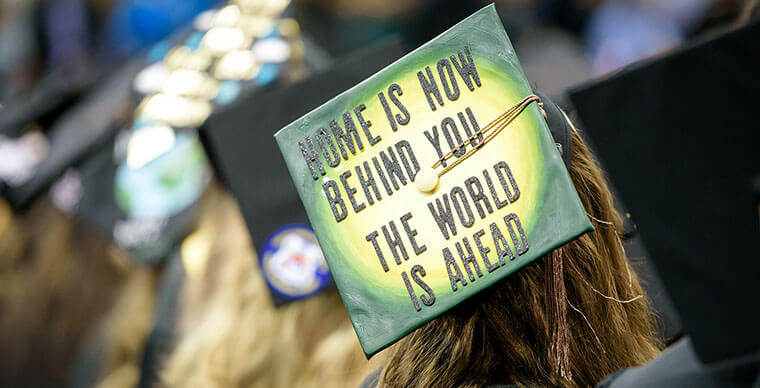 An SPU grad's mortarboard reads "Home is now behind you - the world is ahead."